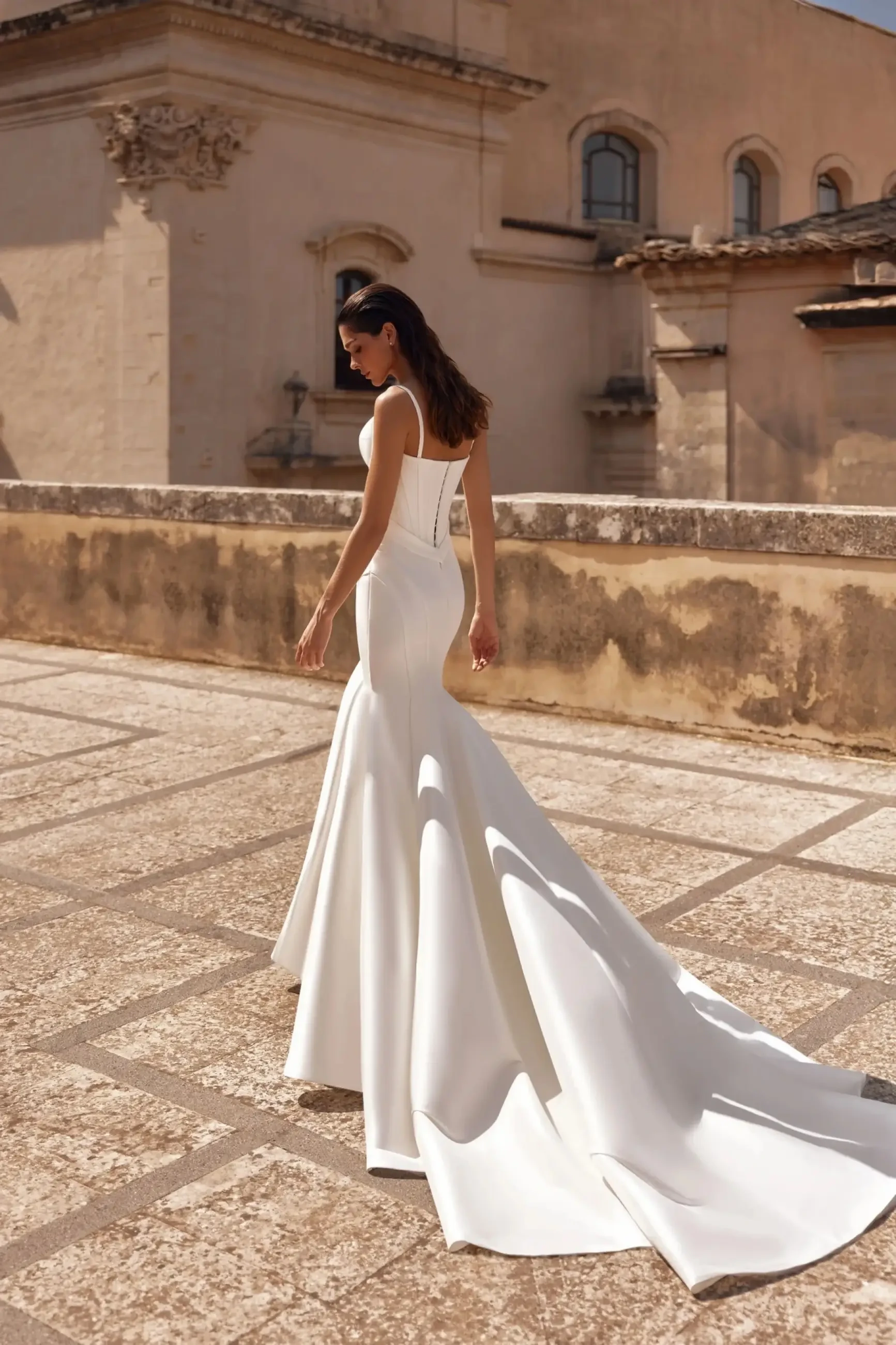 A woman in a sleek, white wedding gown with a long train, standing on a stone terrace.