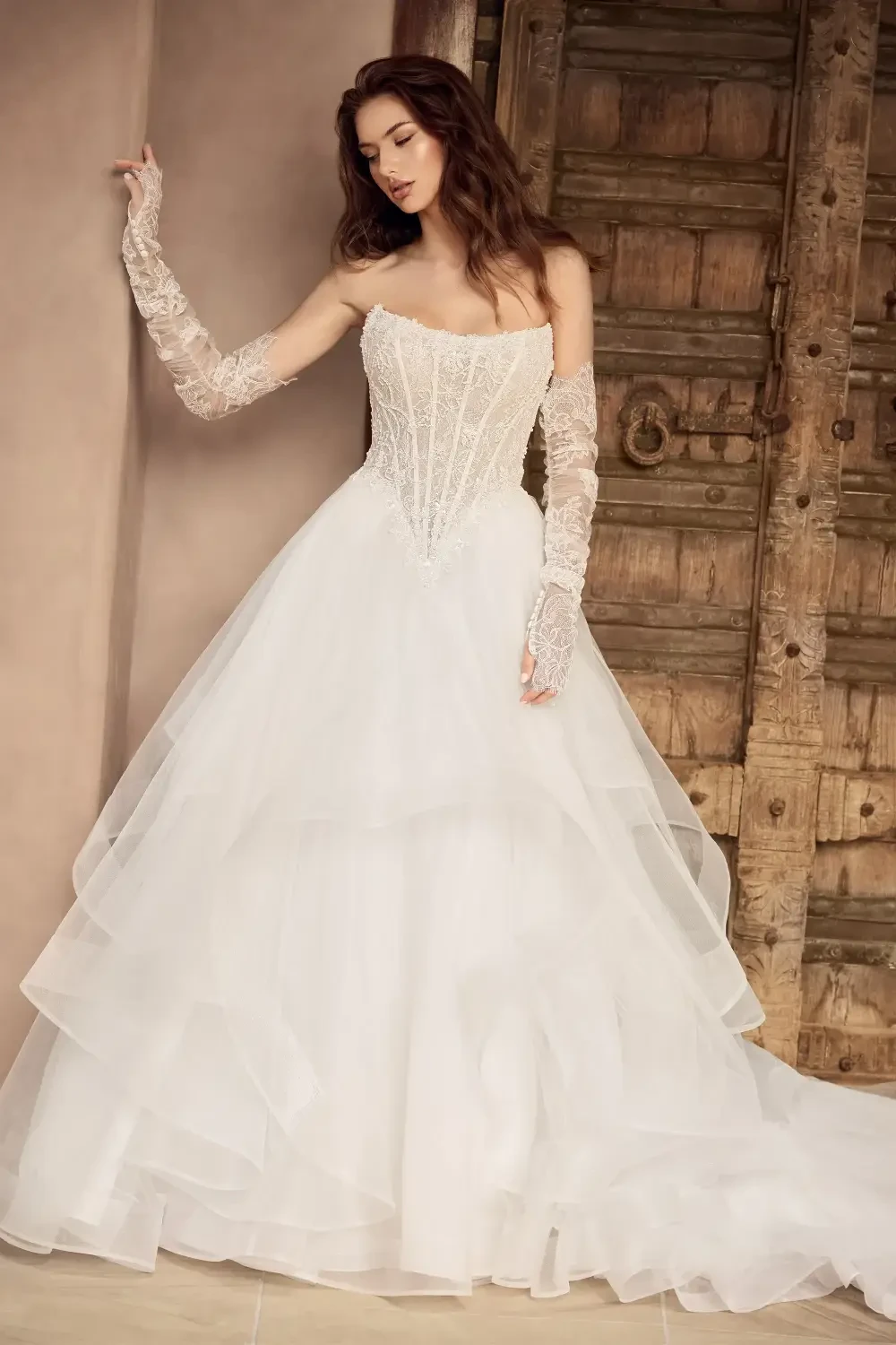 A model wearing a strapless wedding dress with a fitted bodice and layered tulle skirt, accessorized with long, sheer lace gloves, posed in front of a rustic wooden door.
